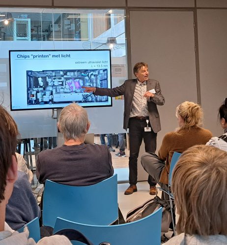 Wim van der Zande, director of ARCNL, gives a lecture to a mixed audience. He points to the screen, which shows a slide titled 'Chips "printen" met licht', which is Dutch for '"Printing" chips with light', and shows an illustration of the inside of a nanolithography machine used to make computer chips.