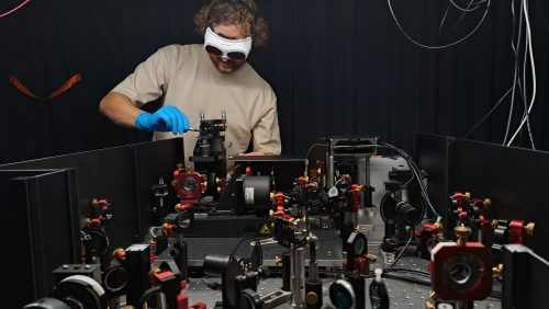Falco Bijloo wears large white optical goggles and a blue glove holding tweezers, with which he adjusts the experimental setup. In front of him lies an optical setup, with many mirrors and other devices. 