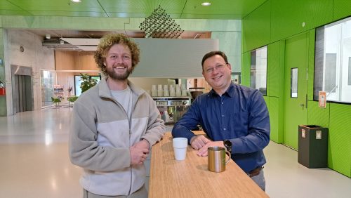 Falco (left) and Femius (right) lean on a high table in front of a coffee machine and smile at the camera.