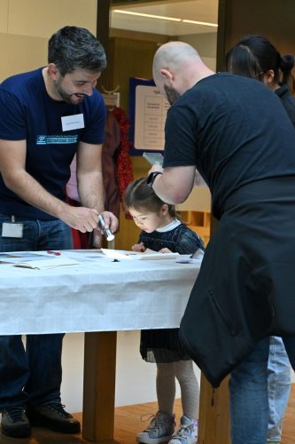 From left to right: An ARCNL researcher shines a flashlight on a piece of chocolate on a table. A young child leans over and looks at the chocolate curiously. Her father points his phone at the chocolate, possibly to take a photo. Her mother stands behind them.