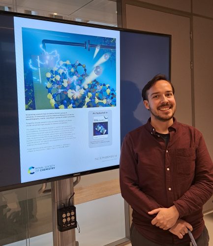 Lukas Hückmann poses smiling in front of a screen displaying the article cover, featuring an image he designed. The image shows a cluster of colorful spheres, electric discharge and a battery.