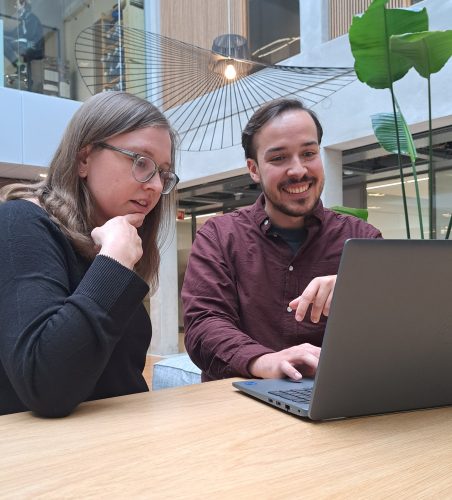 Emilia Olsson (left) and Lukas Hückmann (right) sit at a table looking at a laptop and discussing what they are looking at, which is not visible in the photo.
