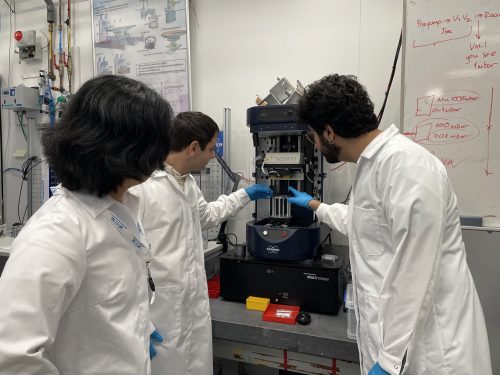 Dennis Kang, Enrico Pedretti and Ozan Sahin place a diamond sample in an experimental setup. They are wearing white lab coats and blue gloves.